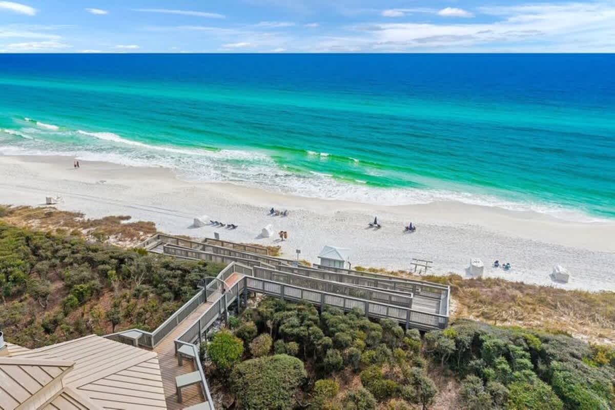 A scenic view from a wooden boardwalk overlooking a wide expanse of white sand beach and the vibrant turquoise ocean with gentle waves, under a bright blue sky.