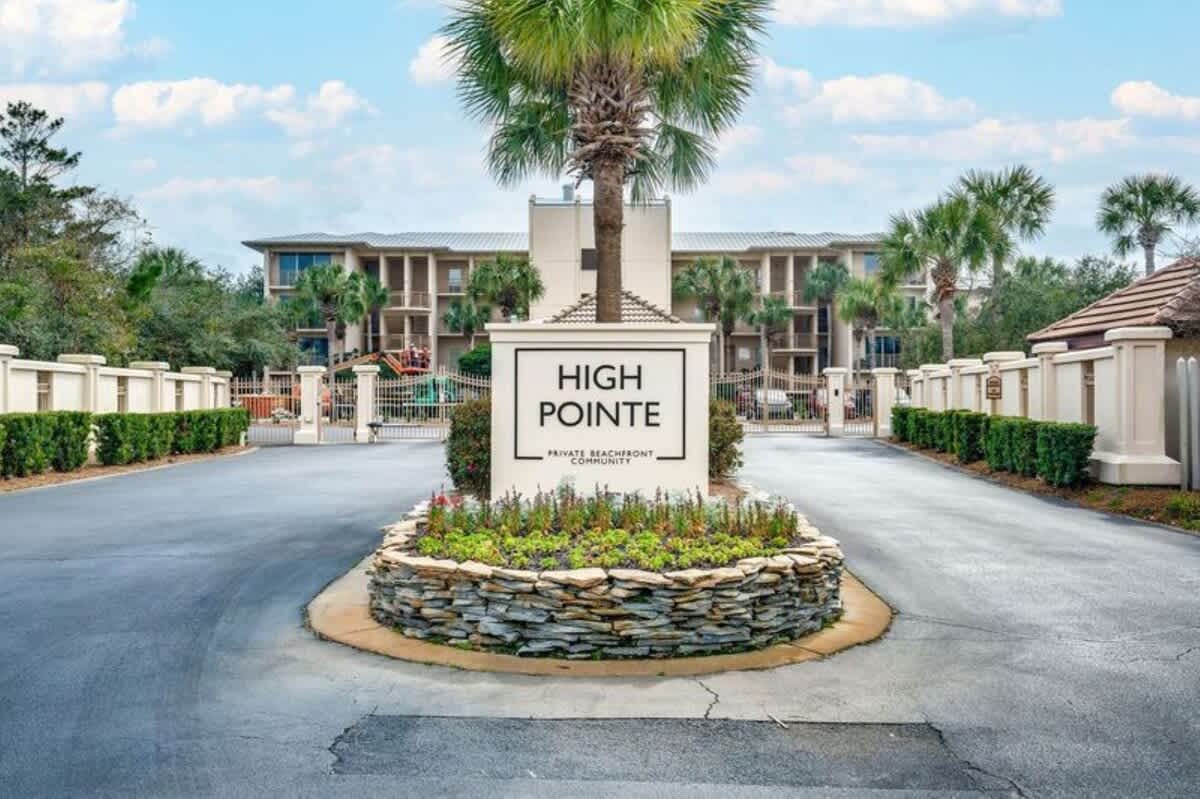 Grand entrance to "High Pointe Private Beachfront Community" with a large white sign, lush landscaping, and palm trees. A paved driveway leads to the gated resort entrance.