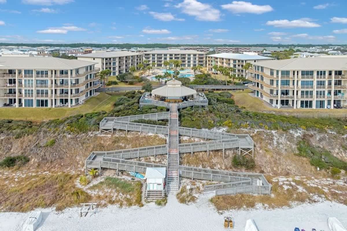 An aerial view centered on a complex wooden boardwalk system leading down to a white sand beach, flanked by multiple large resort buildings and a communal pool area with palm trees.