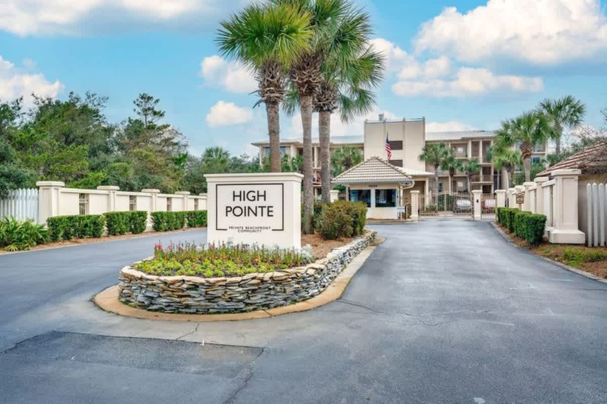 Wide shot of the "High Pointe Private Beachfront Community" entrance, featuring a prominent sign, manicured landscaping, and a guard booth. Palm trees line the paved road leading into the resort.