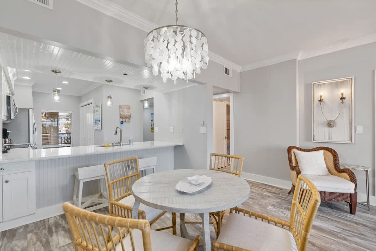Coastal-style dining area with a round light wood table and rattan chairs, featuring a shell-disc chandelier. An adjacent kitchen breakfast bar has white shiplap paneling and pendant lights, with a wicker armchair in the corner.