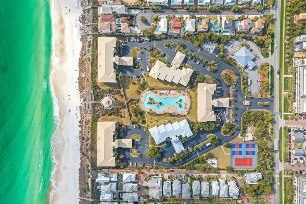 Overhead aerial view of a beachfront resort complex featuring multiple light-colored buildings, a large freeform swimming pool, parking areas, and lush landscaping, all situated alongside a white sand beach and turquoise ocean.