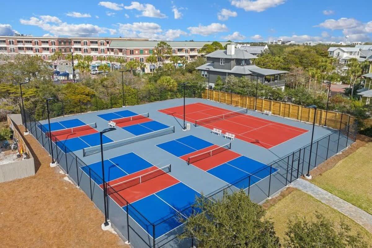 Aerial perspective of a well-maintained outdoor sports complex featuring one full-sized red tennis court and multiple smaller blue and red pickleball courts, enclosed by fencing and surrounded by green foliage.