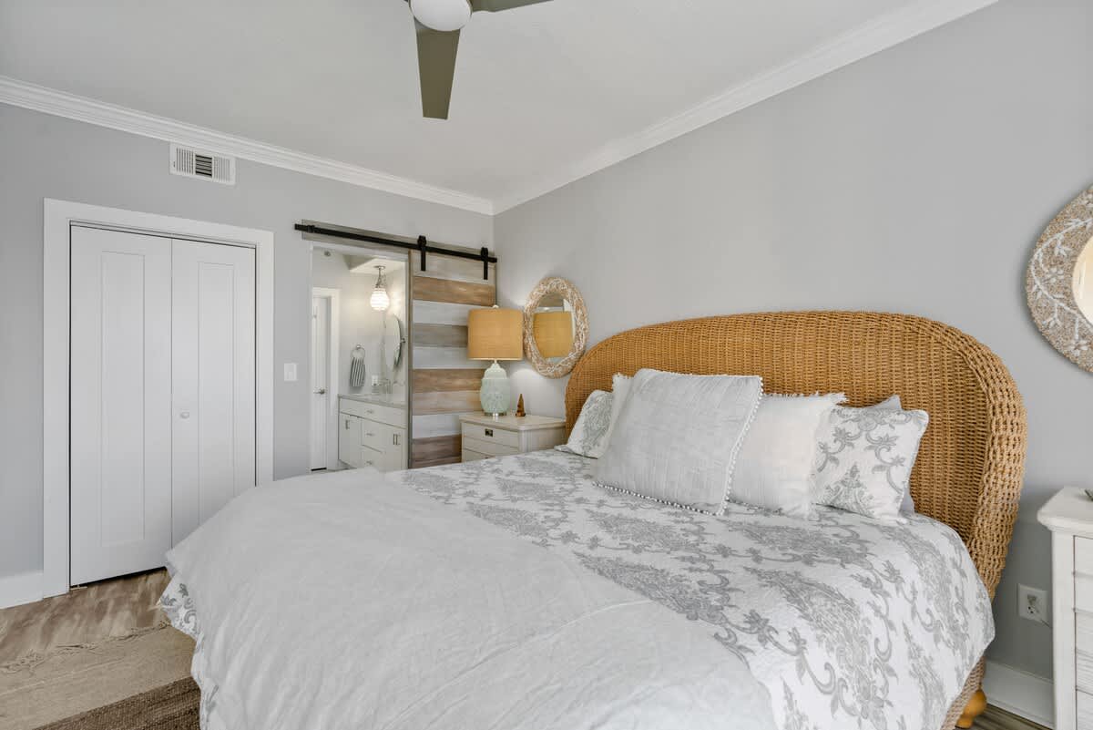 Coastal-themed bedroom with a king bed featuring a wicker headboard and light bedding, complemented by a sliding barn door revealing a modern en-suite bathroom.