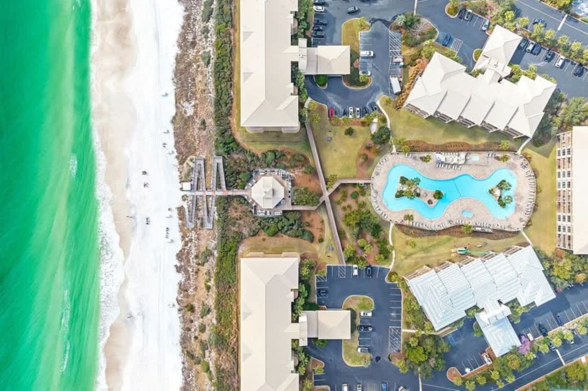 High-angle aerial view of a resort property nestled along a pristine white sand beach with emerald green ocean waters, featuring several large buildings, a winding communal pool, and wooden boardwalks leading to the beach.