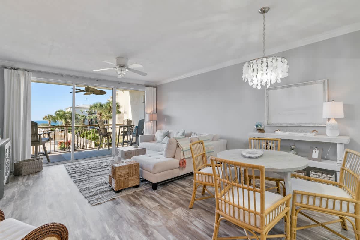 Open-concept living and dining area featuring a light sectional sofa, a round dining table with rattan chairs, and a large sliding glass door leading to a balcony with views of a pool and palm trees.