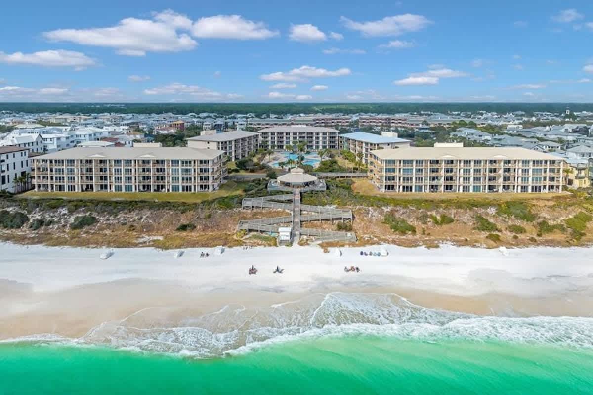 A broad aerial view of several large, multi-story resort buildings with light roofs, positioned along a white sand beach with turquoise ocean waves, connected by a prominent wooden boardwalk system.