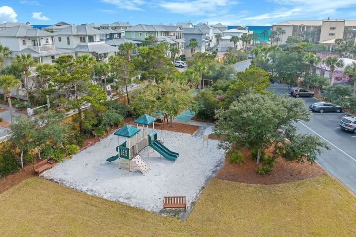 An aerial view of a sandy playground featuring a green and tan playset with slides and climbing elements, surrounded by lush green trees and residential houses, with the ocean visible in the distance.