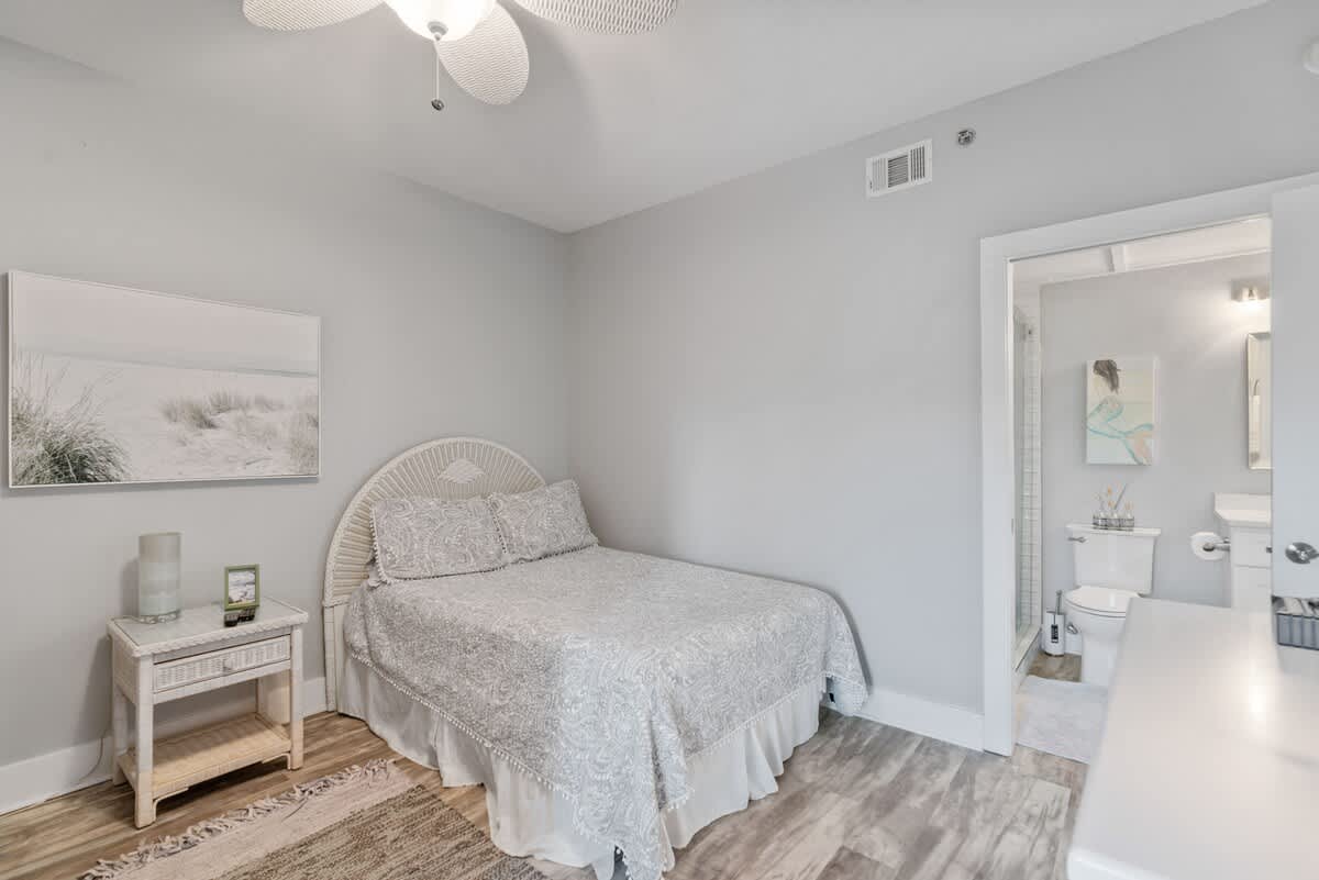 Cozy bedroom corner showcasing a queen bed with a white wicker headboard and a matching white nightstand with a lamp. Light wood-look flooring and a textured rug complete the inviting space.