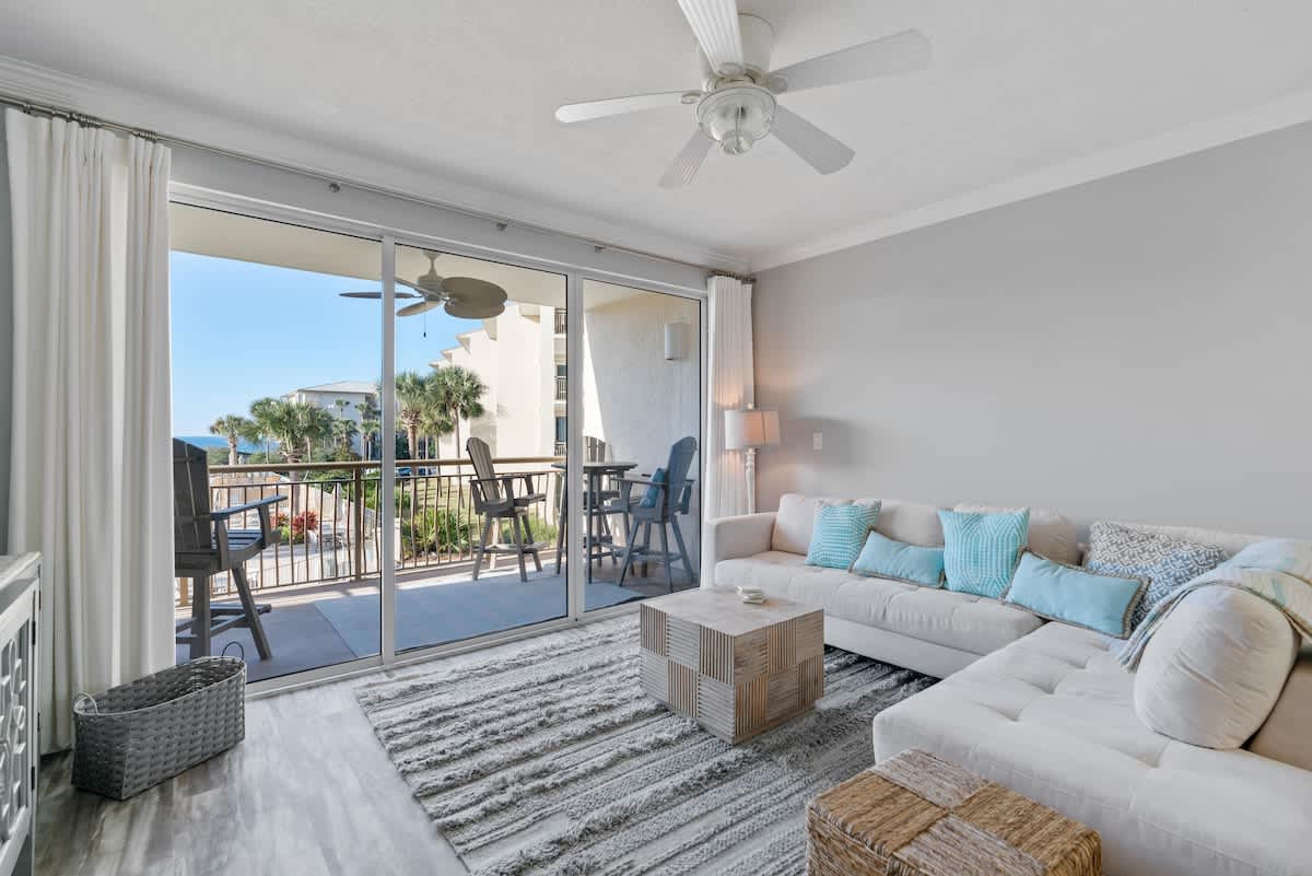Inviting living room with a light-colored sectional sofa and a textured rug, offering a clear view through sliding glass doors to a balcony. The balcony overlooks a resort pool area with palm trees and a glimpse of the ocean.