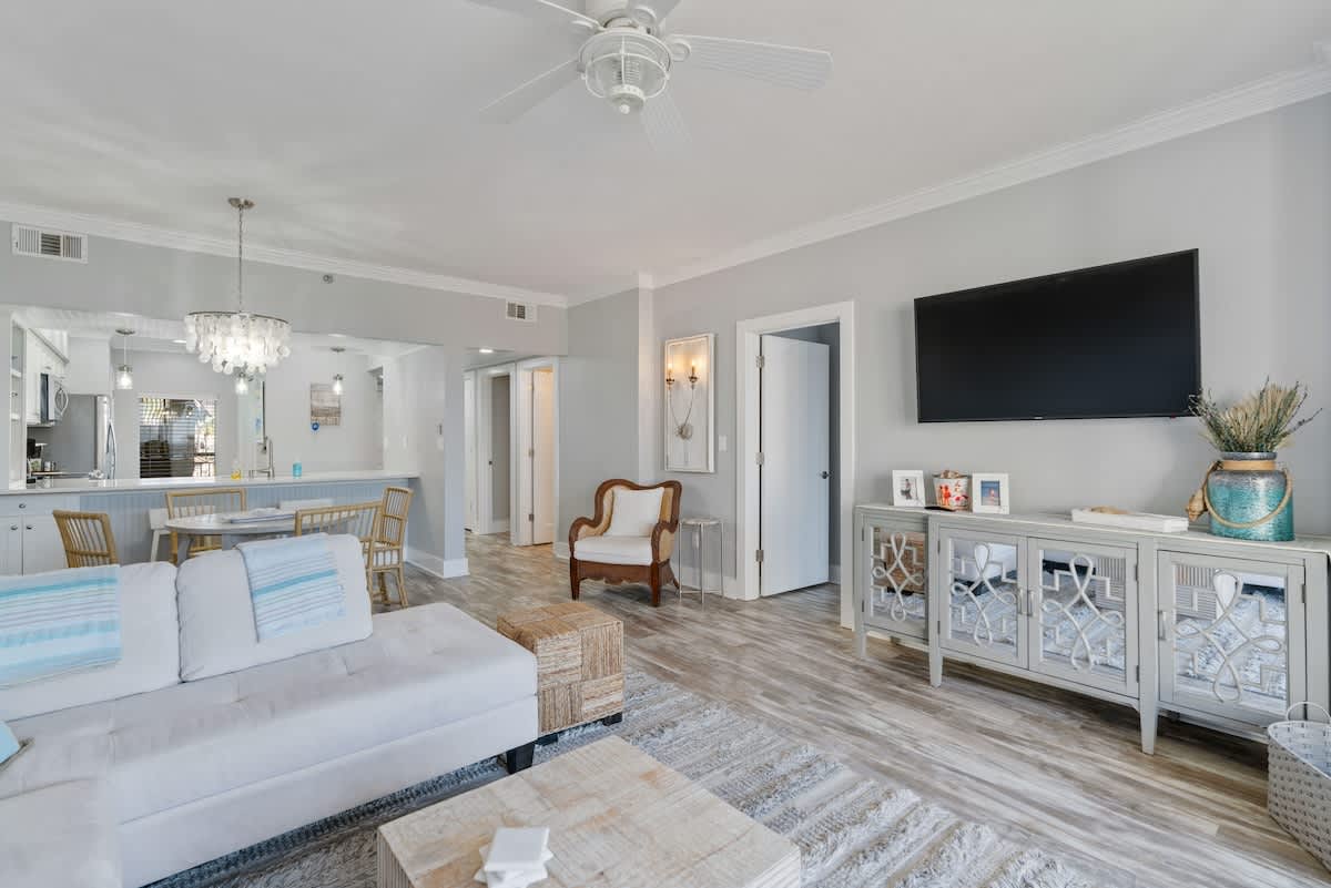Open-concept living room with a large L-shaped sofa and a textured rug, leading into a dining area with a round table and a modern chandelier. A glimpse of the kitchen with white cabinetry is visible beyond.
