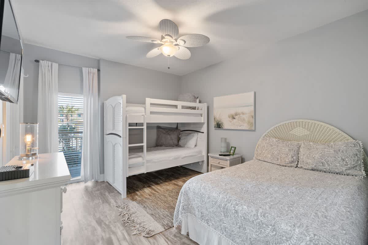 Bright bedroom with light gray walls and wood-look flooring, featuring a queen bed with a white wicker headboard and a white bunk bed. A ceiling fan with leaf-shaped blades adds a tropical touch.