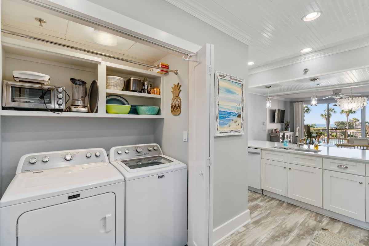 Compact laundry area featuring a white top-load washer and dryer, with open shelving above holding kitchen appliances and dishes, located adjacent to the modern kitchen.