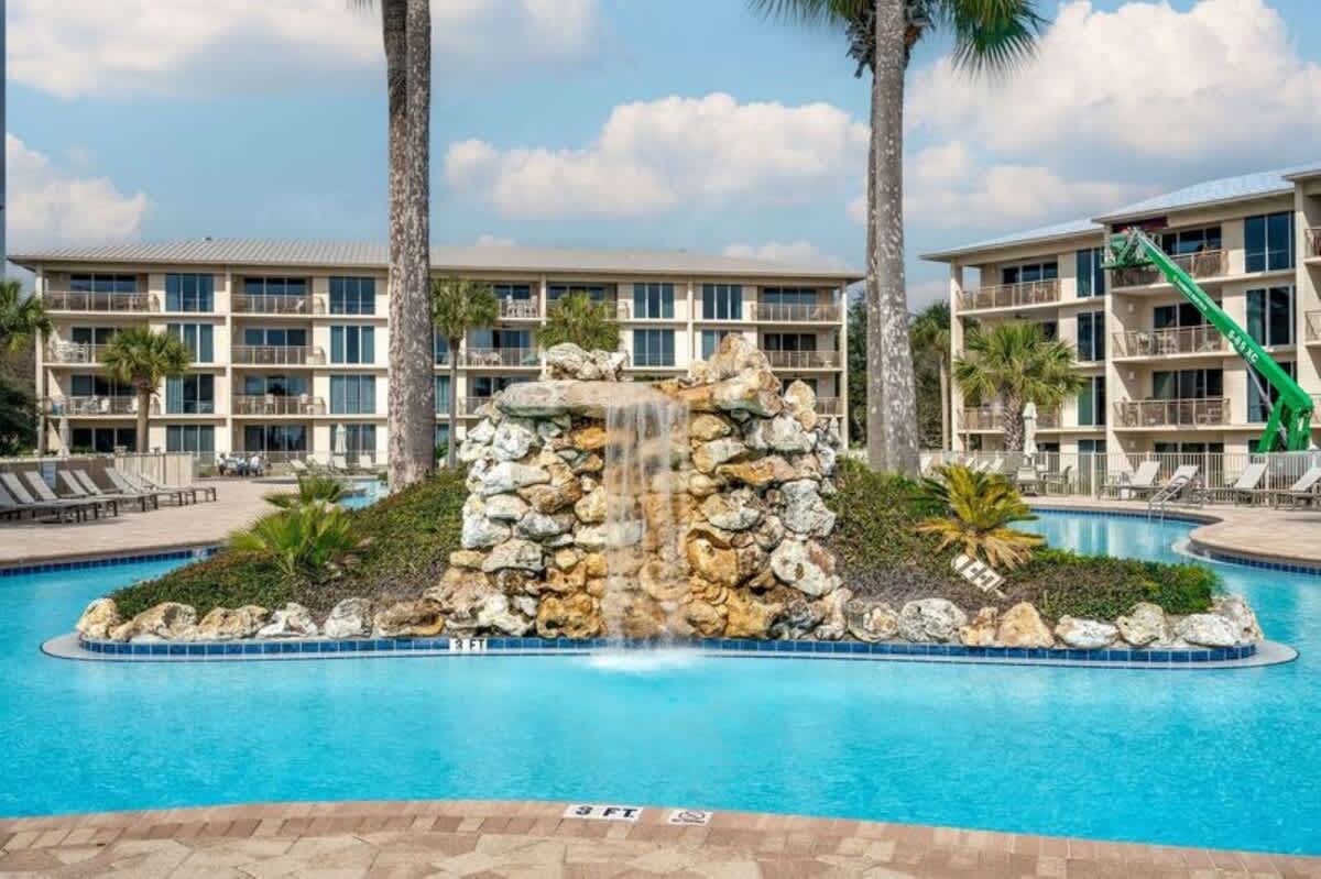 Detailed view of a decorative rock waterfall feature cascading into the communal swimming pool, surrounded by lush tropical plants and tall palm trees, with resort buildings in the background.