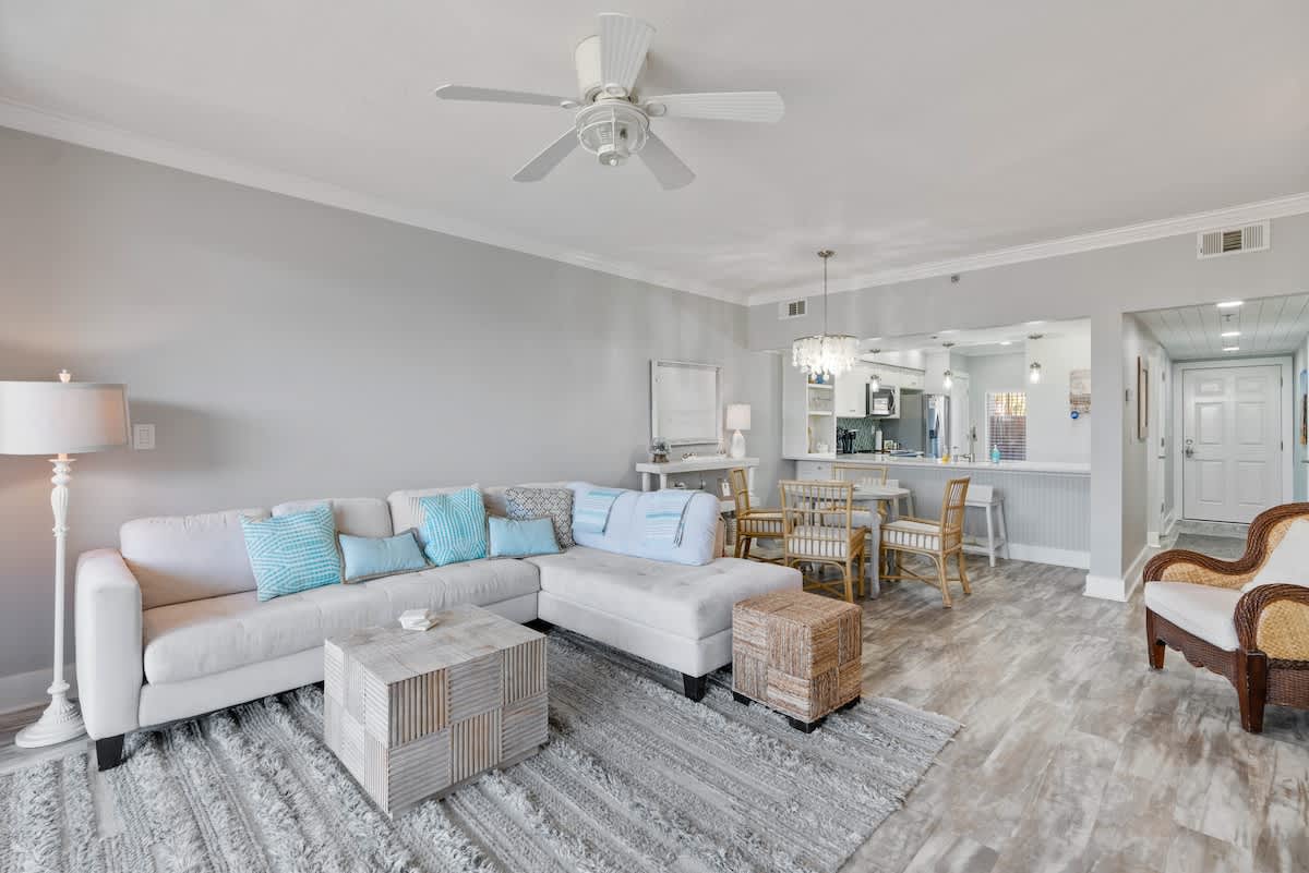 Spacious living room with a light sectional sofa and a woven ottoman, facing a dining area with a round table and a breakfast bar. The room features light gray walls, wood-look flooring, and a ceiling fan.