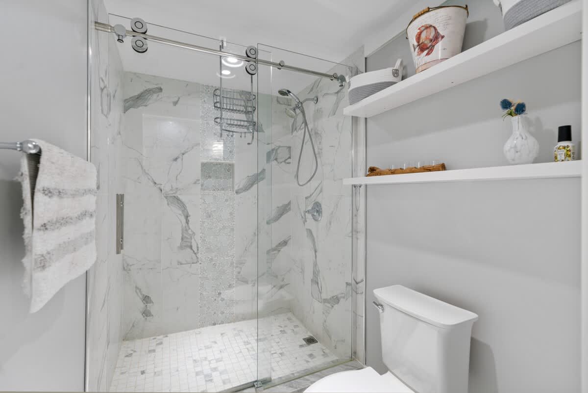 Spacious walk-in shower with clear glass sliding doors, white marble subway tiles, and a mosaic accent strip. A white toilet is visible next to the shower, with open shelving above.