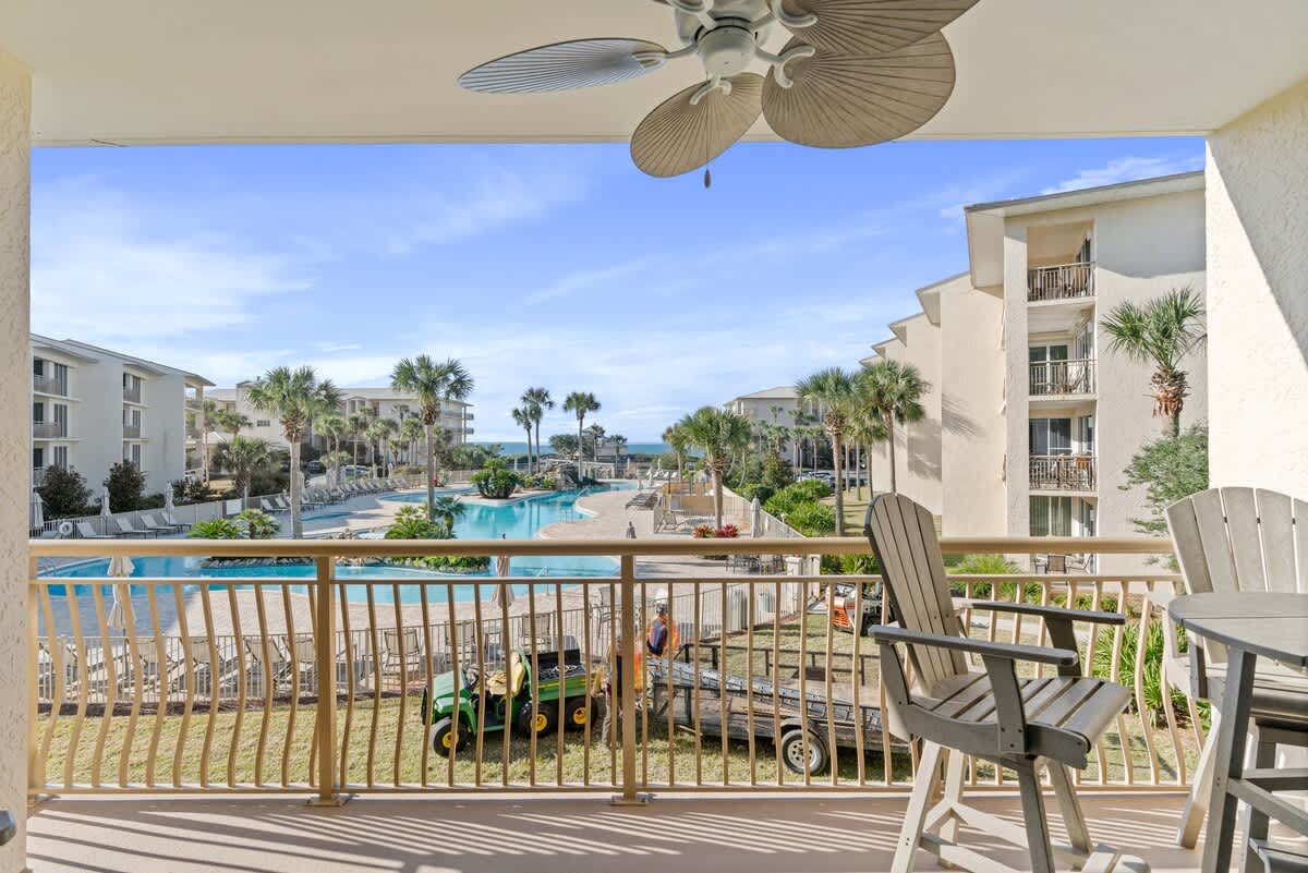 Expansive balcony overlooking a large communal pool area surrounded by palm trees and resort buildings, with a glimpse of the ocean in the distance. Outdoor seating is visible.