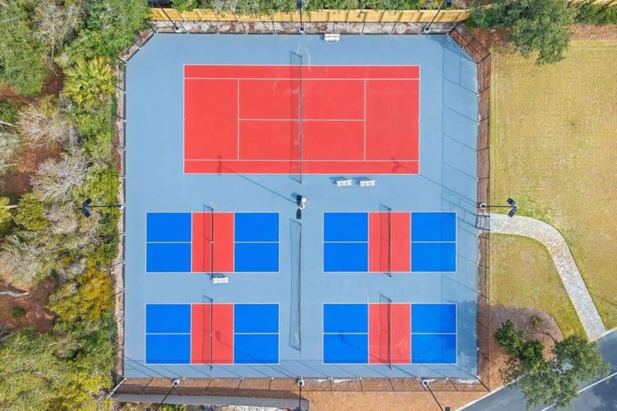 Direct overhead aerial shot showcasing a vibrant outdoor sports area with a large red tennis court at the top and four smaller blue and red pickleball courts below, all clearly marked and surrounded by fencing.