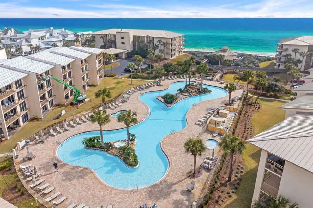 Expansive aerial view of a large, freeform communal swimming pool with multiple lounge chairs and palm trees, surrounded by resort buildings, with the vibrant turquoise ocean visible in the background under a blue sky.