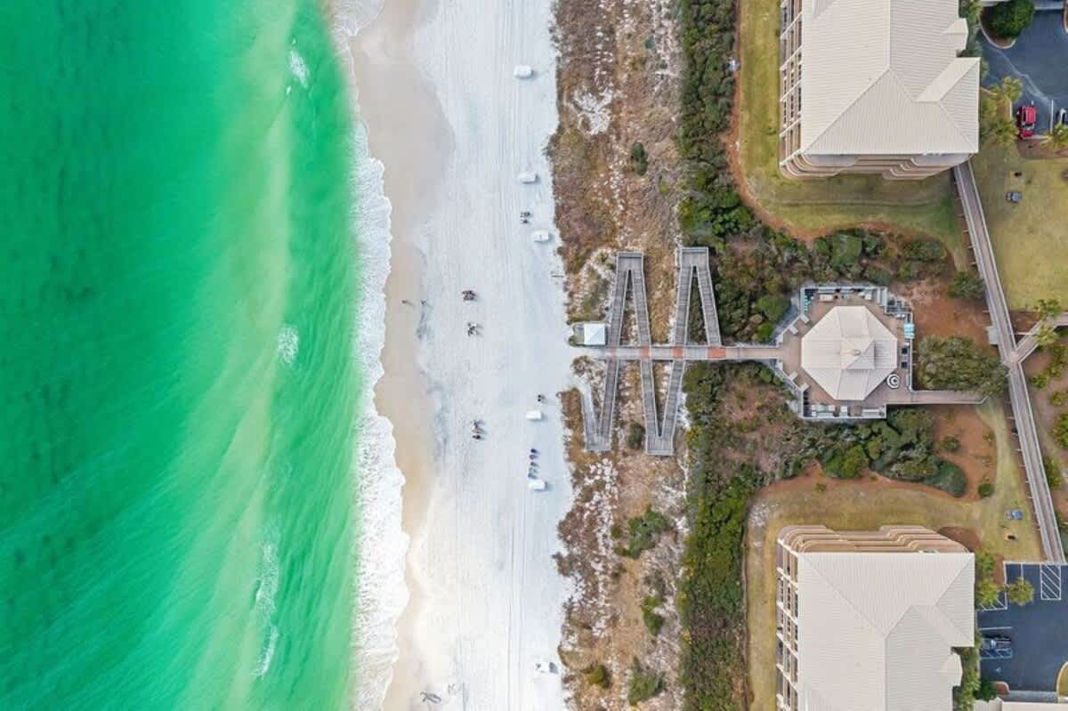 An aerial, top-down view of a vibrant turquoise ocean meeting a wide white sand beach, with a wooden boardwalk and a circular building connecting to multi-story resort buildings on the right.