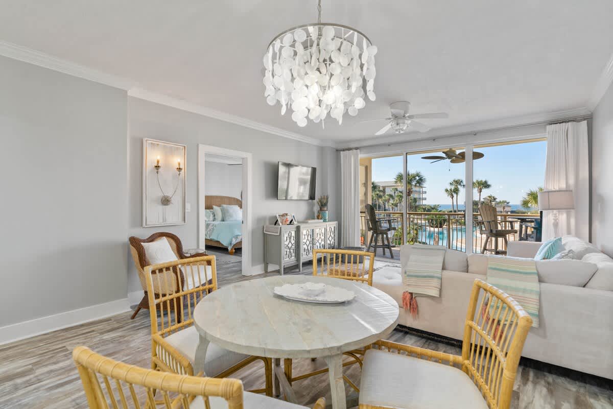 Spacious living and dining area with a light sectional sofa, round dining table, and a large sliding glass door opening to a balcony with views of the pool and ocean. A bedroom entrance is visible to the left.