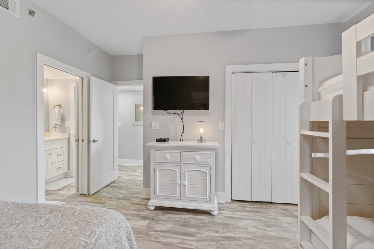 View of the bedroom showing a wall-mounted TV above a white dresser, with a bunk bed partially visible on the right. A doorway leads to an adjacent bathroom, revealing a vanity inside.