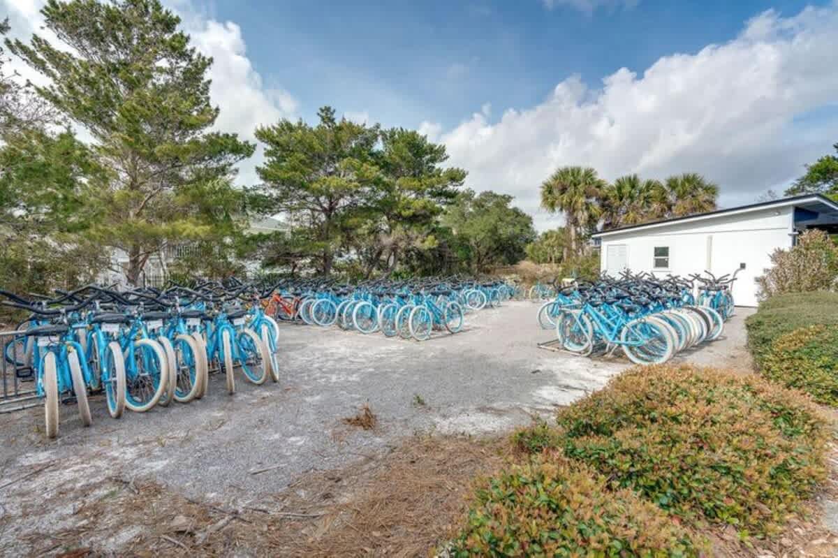 A large outdoor area filled with rows of light blue bicycles parked on racks, surrounded by trees and a small white building under a partly cloudy sky.