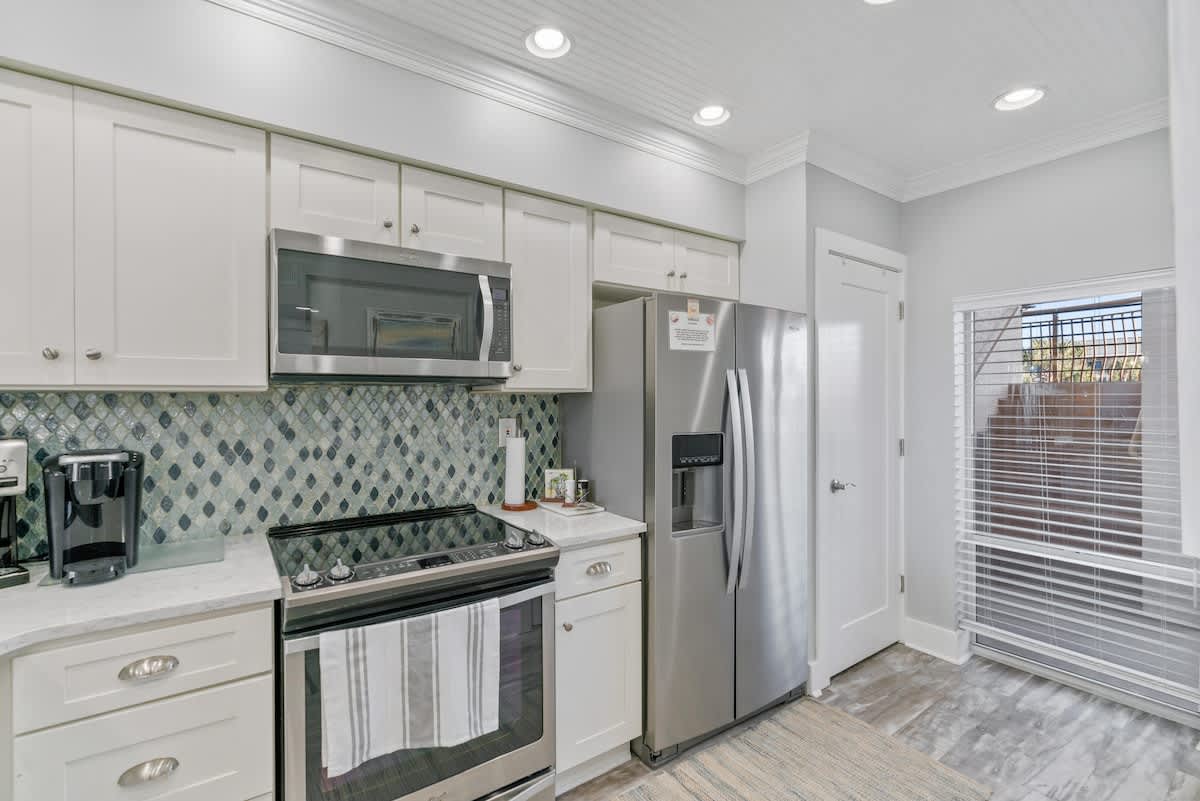 Well-equipped kitchen featuring white shaker cabinets, stainless steel appliances including a microwave, range, and side-by-side refrigerator. The backsplash is a unique blue and green mosaic tile, complementing the light countertops.
