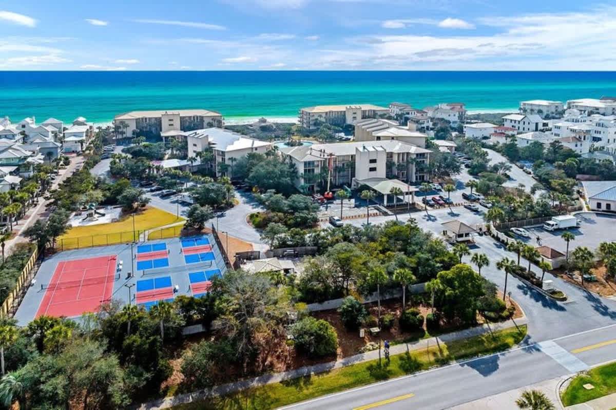 Expansive aerial view of a coastal resort community with several multi-story buildings, lush green spaces, roads, and a vibrant red and blue sports court, all set against the backdrop of a bright blue sky and turquoise ocean.