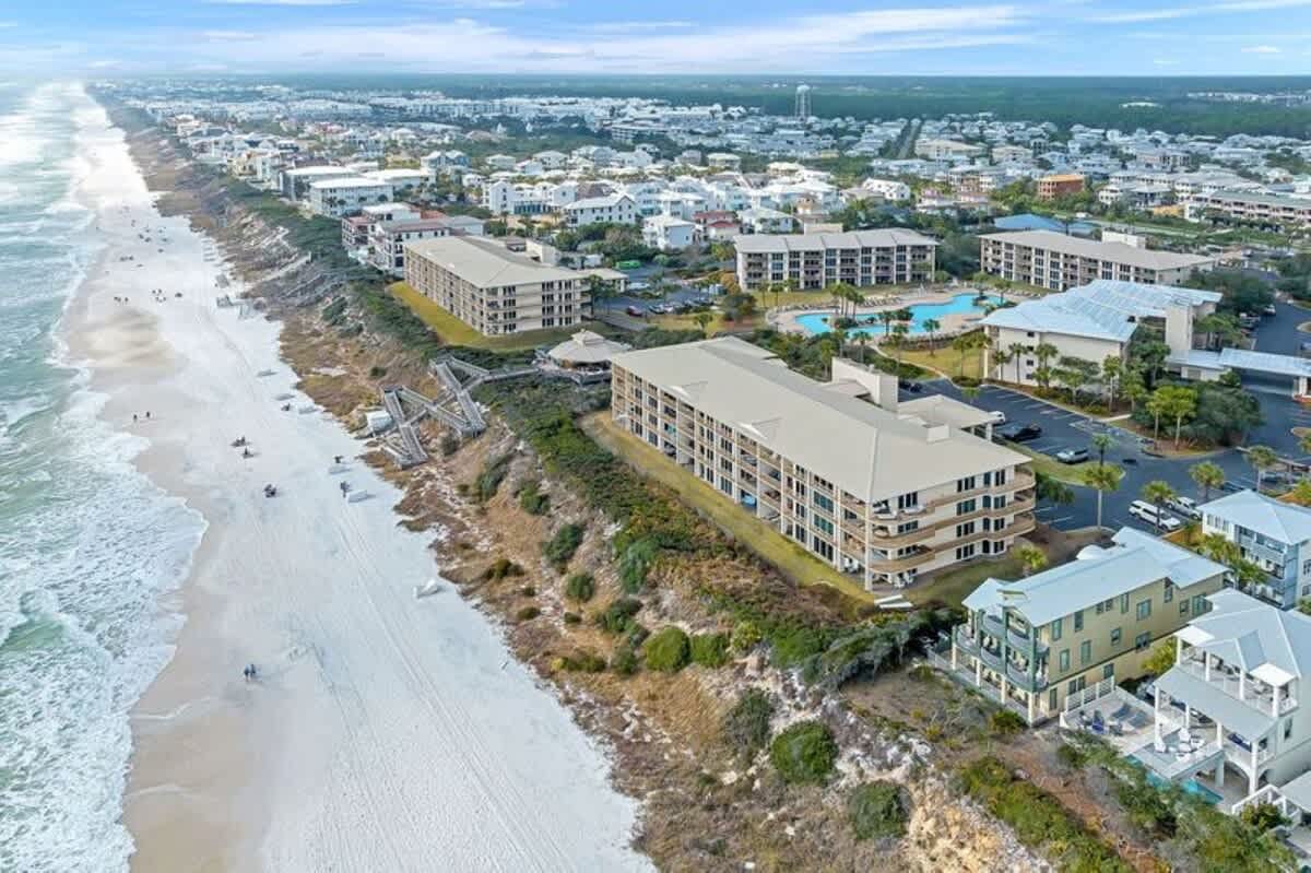 A wide aerial view of a long stretch of white sand beach and turquoise ocean, bordered by multiple large resort buildings with light roofs and lush coastal vegetation, extending into the distance under a partly cloudy sky.