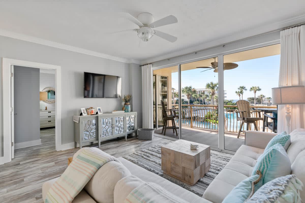Cozy living room with a light sectional sofa and a wooden coffee table, featuring a large flat-screen TV mounted above a decorative console. Sliding glass doors open to a balcony with views of a pool and lush palm trees.