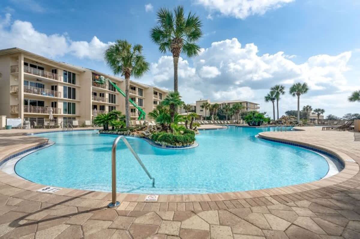 Close-up view of a section of the large communal swimming pool, featuring steps leading into the clear blue water, a metal handrail, and a patterned paver deck, with palm trees and resort buildings in the background.