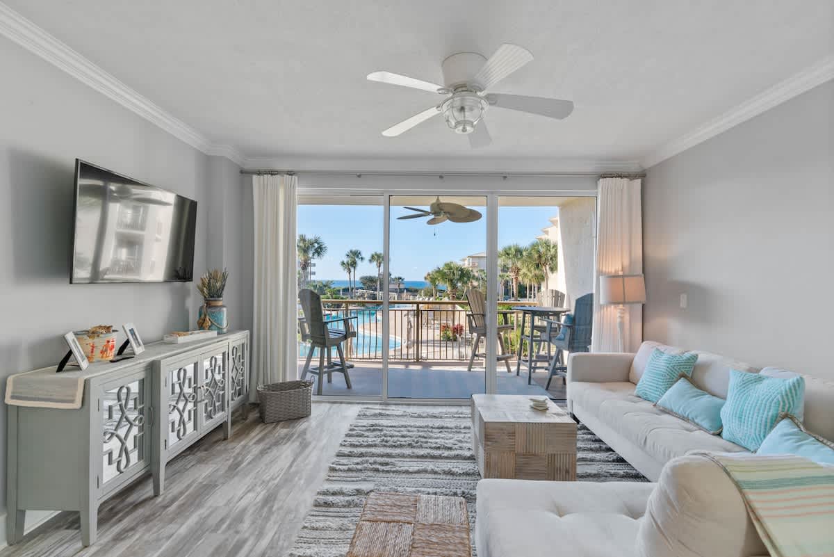 Modern living room featuring a light gray sectional sofa with blue accent pillows, a textured area rug, and a square wooden coffee table. Sliding glass doors lead to a balcony with views of a pool, palm trees, and the ocean.
