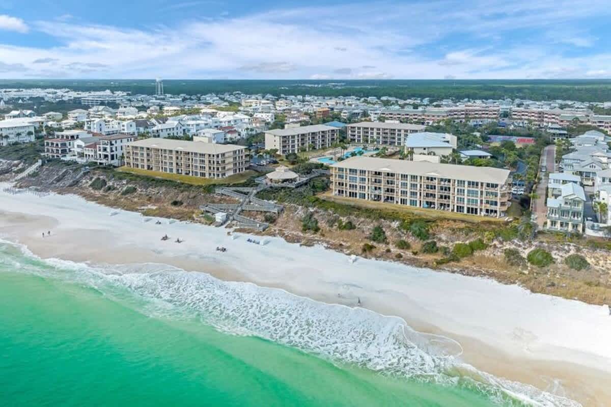 An aerial view showcasing multiple large resort buildings with light-colored roofs situated on a sandy bluff overlooking a wide white sand beach and a vibrant turquoise ocean under a blue sky.