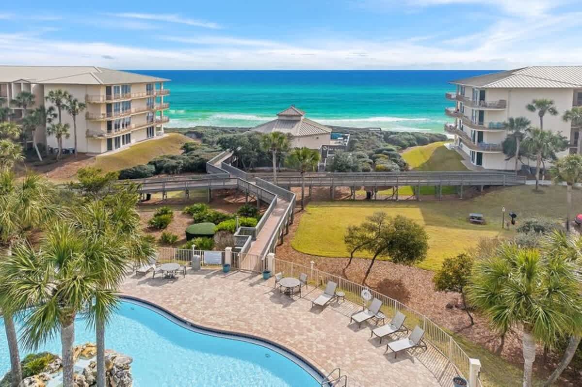 Scenic view from the pool area towards the ocean, showcasing elevated wooden boardwalks leading to a beachside pavilion, flanked by resort buildings and lush greenery, with the bright blue sea in the distance.