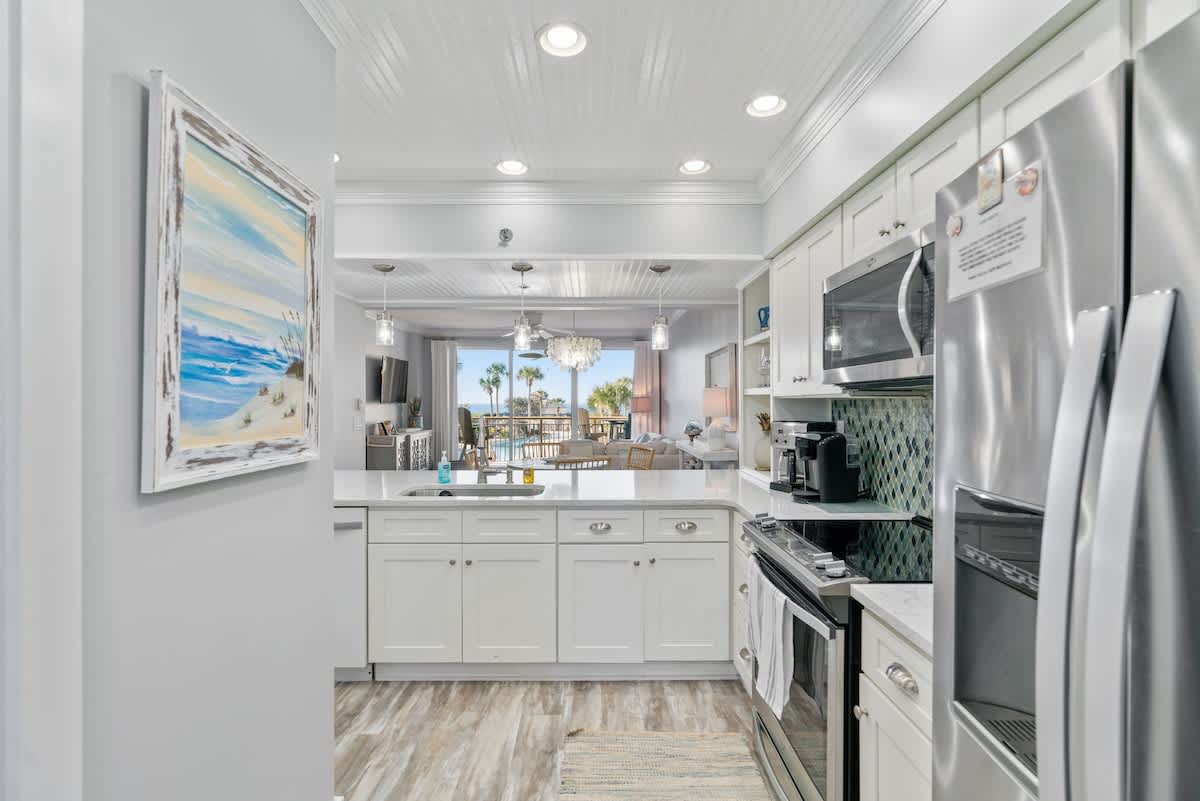 Contemporary kitchen with white cabinetry, light countertops, and a distinctive blue and green mosaic tile backsplash, equipped with stainless steel appliances. The open layout provides a direct line of sight through the living room to the outdoor balcony.