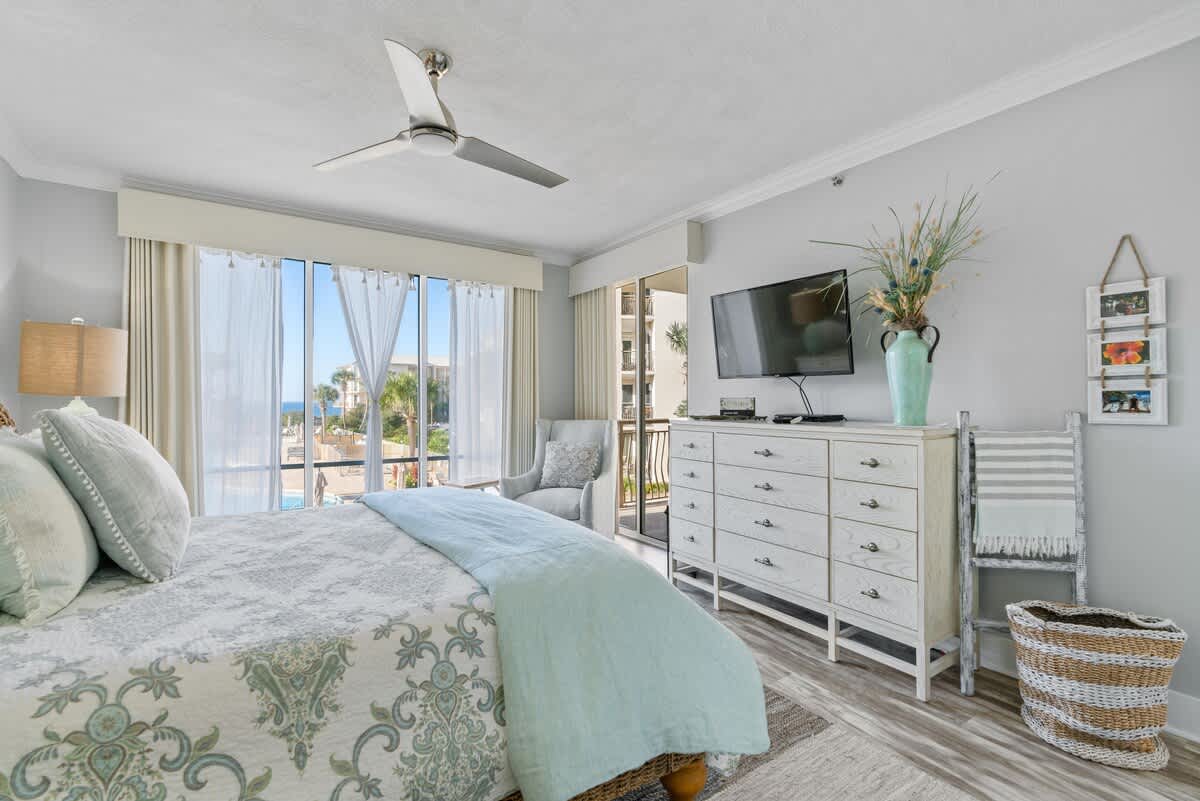 Spacious bedroom featuring a white dresser with a mounted flat-screen TV, a comfortable armchair, and a large window providing access to a balcony with outdoor views.