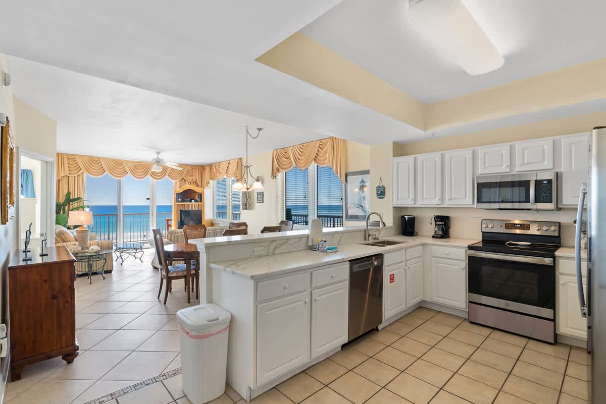 An open-concept kitchen with white cabinets and stainless steel appliances overlooks the dining and living areas, which feature large windows with ocean views, all on light tile flooring.