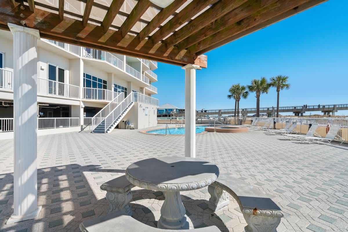 A concrete picnic table with benches sits under a wooden pergola on a patterned paver deck, adjacent to the communal pool area and the high-rise building, with the ocean visible in the distance.