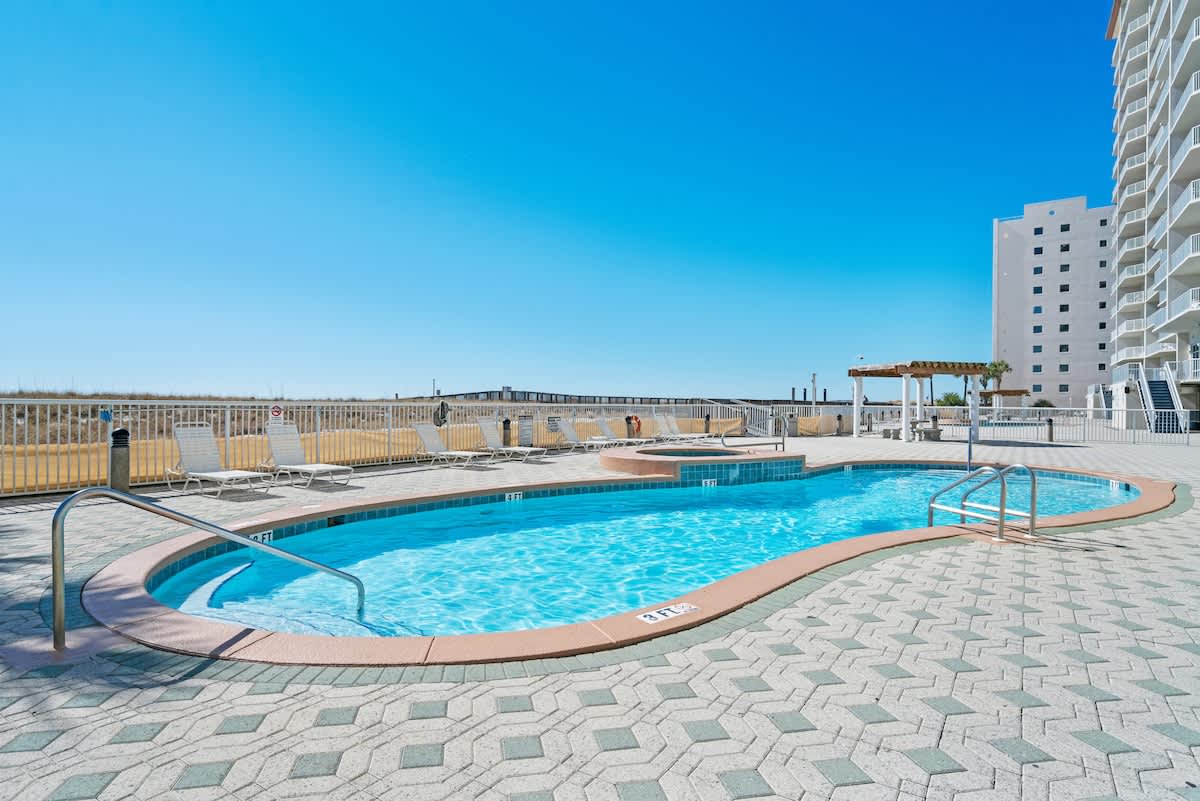 A kidney-shaped communal swimming pool with blue water and a brown coping is surrounded by a patterned paver deck, featuring lounge chairs and a hot tub under a clear blue sky.