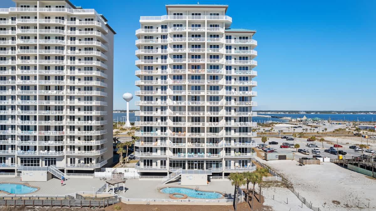 Close aerial view of two identical white condominium buildings with multiple balconies, featuring two outdoor swimming pools in the foreground and a bay with boats visible in the distant background.