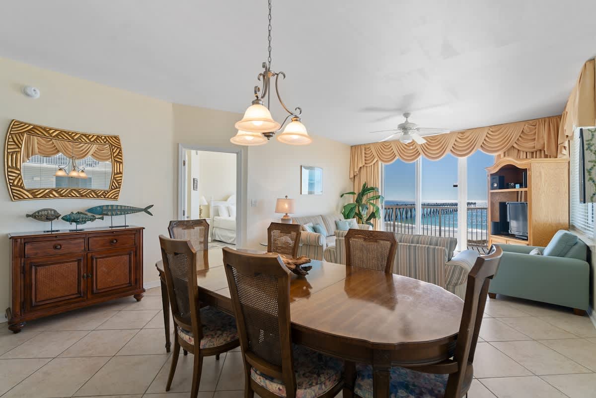 An oval wooden dining table with six chairs featuring woven backs and floral cushions sits on light tile floors, adjacent to a living area with ocean views through large windows.