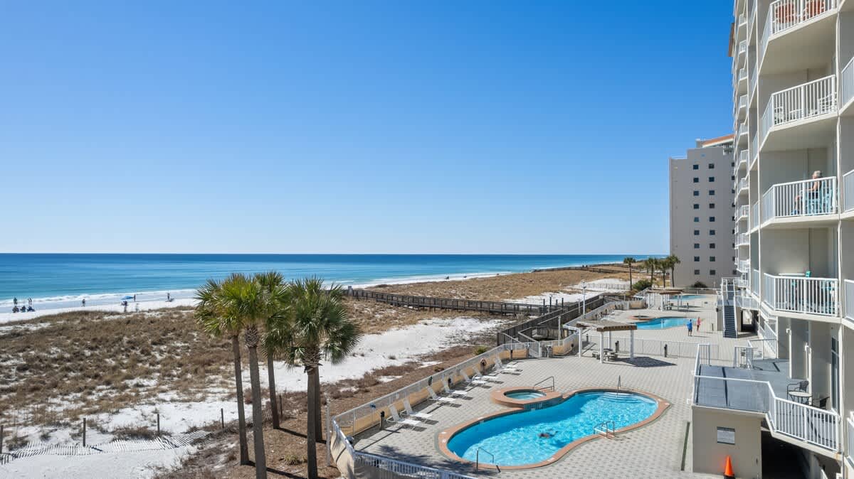 View from an upper-floor balcony overlooking a communal swimming pool area, a wooden boardwalk traversing natural dunes, a wide white sand beach, and the expansive blue ocean under a clear sky.