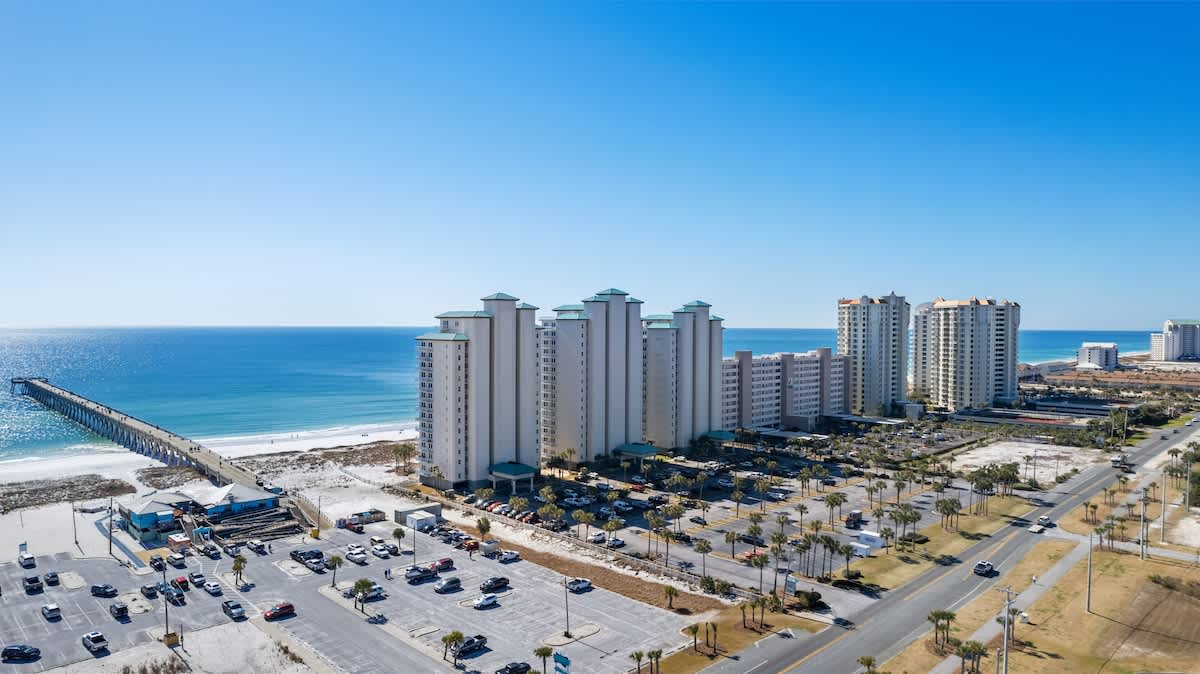 Wide aerial view showcasing multiple white condominium buildings with teal roofs along a long white sand beach, with a prominent pier extending into the turquoise ocean and a coastal road.