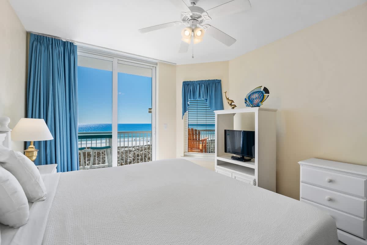 A corner view of Bedroom 1, highlighting the white king bed, a white dresser, and the sliding glass door to the balcony, offering clear ocean and pier views.