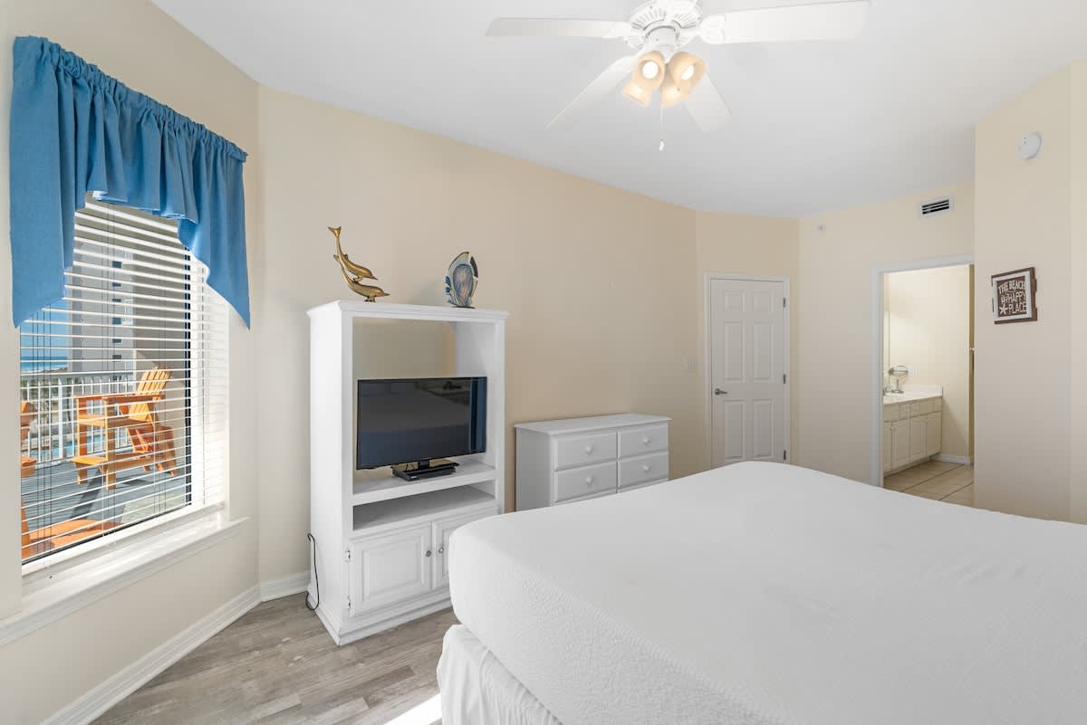 Bedroom 1 features a white king bed, a white entertainment center, and a window with blinds, with a doorway leading to an en-suite bathroom, all on light wood-look flooring.