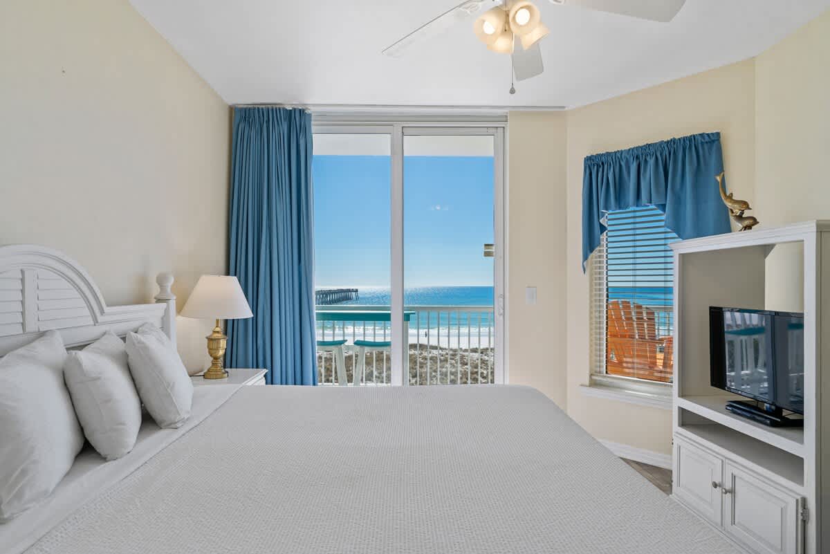 View from the foot of the white king bed in Bedroom 1, showing a white entertainment stand with a TV, a window with blue valance and blinds, and a sliding glass door to the ocean-view balcony.