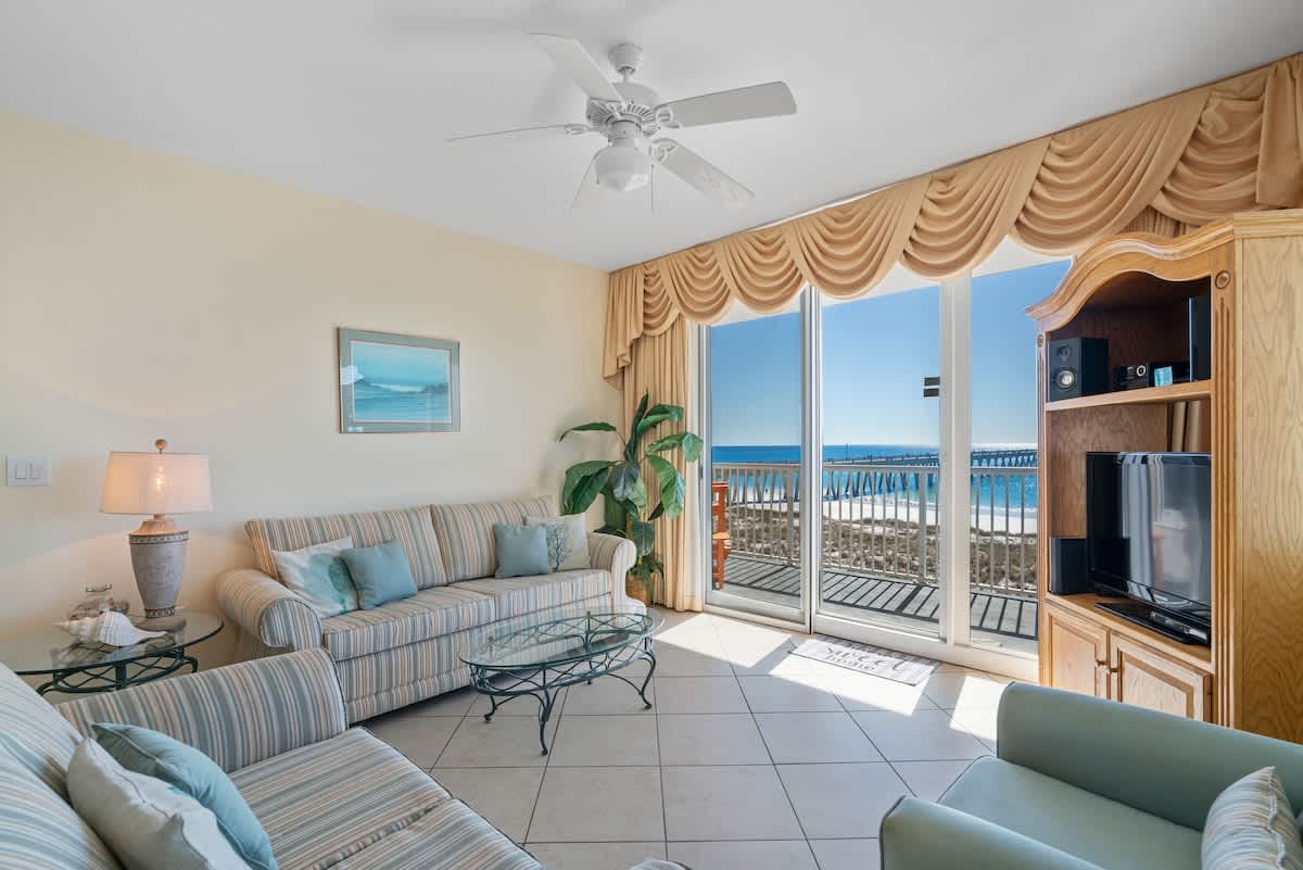 Inviting living room with light tile floors, two striped sofas, a glass coffee table, and a large sliding glass door providing abundant natural light and a stunning view of the ocean and distant pier.