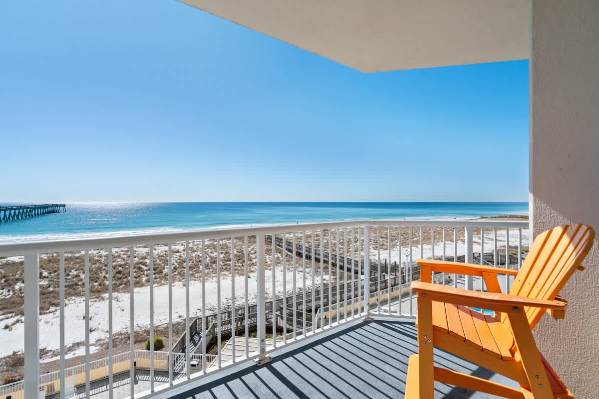 A bright orange Adirondack chair sits on a private balcony with a gray floor, offering expansive views of a white sand beach, the turquoise ocean, and a distant pier under a clear blue sky.