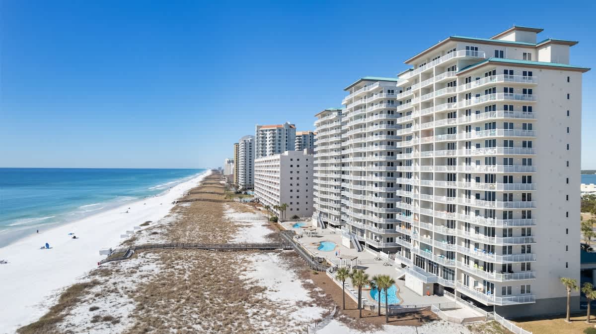 Aerial view of a tall, white condominium building with teal roofs and numerous balconies, situated directly on a white sand beach with two outdoor swimming pools and a boardwalk leading to the ocean.